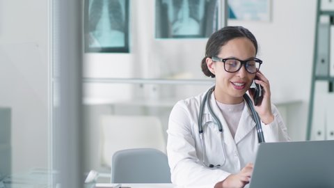 Young mixed-raced female doctor in lab coat, glasses and stethoscope over her neck using laptop, smiling and speaking on mobile phone at desk during workday in clinic
