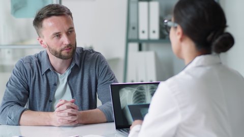 Handsome Caucasian man sitting at table in medical office, looking at x-ray scan of lunges and discussing diagnosis with female physician during consultation