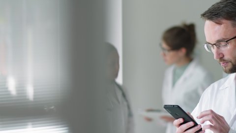 Chest-up shot of Caucasian male doctor in lab coat and glasses standing in medical office and scrolling on smartphone screen while his female colleagues speaking in background