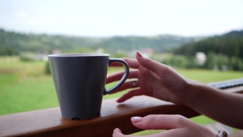 girl picks up a ceramic cup with coffee tea, which stands on the railing of the balcony, against the backdrop of mountains and forest