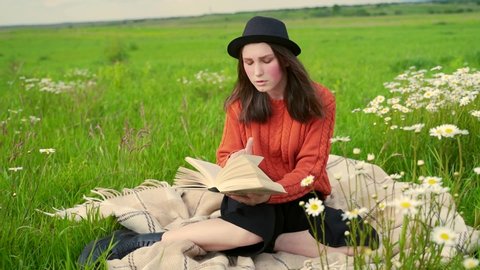 Young cute girl with red cheeks and freckles holding book in her hands. Teenager in a black hat and red sweater sitting in the park and reading book. 