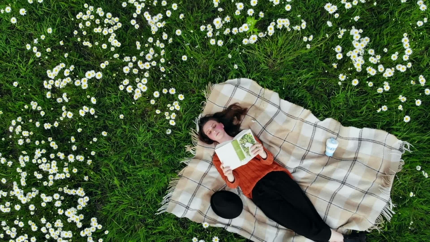 Young teenager with rosy cheeks freckles reading book on lawn on sunny day. Beautiful cute stylish girl with brown hair studying outside in park nature. Children lying with plaid on grass. Aerial view
