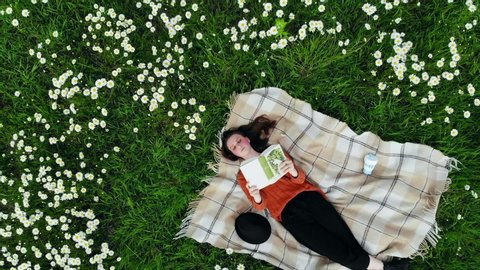 Young teenager with rosy cheeks freckles reading book on lawn on sunny day. Beautiful cute stylish girl with brown hair studying outside in park nature. Children lying with plaid on grass. Aerial view