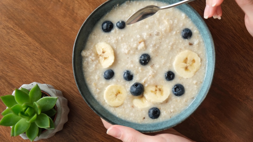 Eating oatmeal porridge with banana and blueberries. Woman's hands stirring breakfast oats in bowl Royalty-Free Stock Footage #1053966029