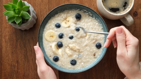 Eating oatmeal porridge with banana and blueberries. Woman's hands stirring breakfast oats in bowl