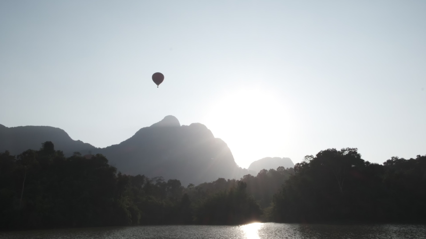 Silhouette of hot air balloon flying above forest behind lake and infant of mountain with sun 