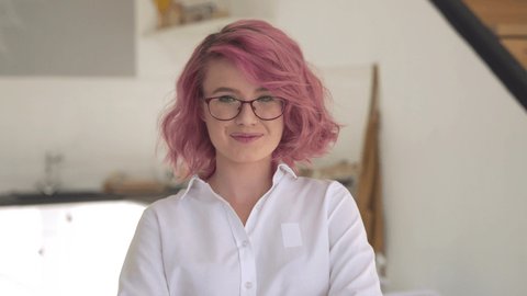 Happy young adult hipster gen z teen girl smiling face with pink hair wearing glasses looking at camera stand in modern kitchen interior. Female small business cafe shop owner entrepreneur portrait.