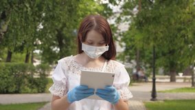 healthy girl is walking in protective mask and medical gloves working with tablet in city park. COVID-19. danger of virus. young business woman working remotely on street. Safe and social distance.