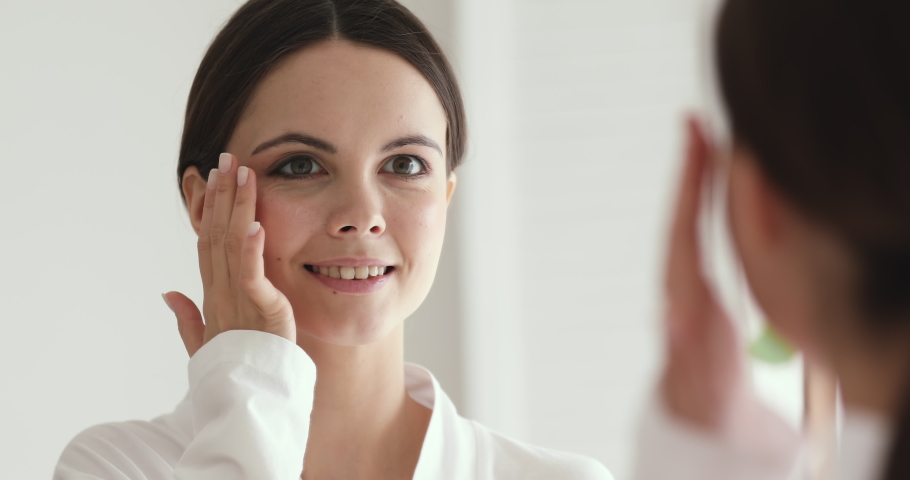 Close up head shot young happy pretty woman in white bathrobe looking in mirror, applying moisturizing cream or nourishing lotion on under eyes area, preventing wrinkles appearance, skincare routine. | Shutterstock HD Video #1053977810