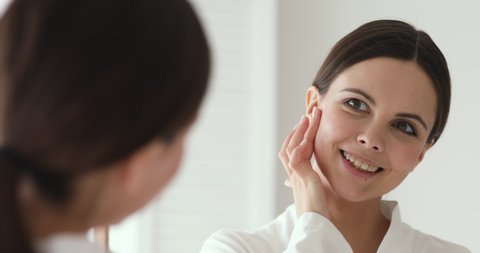 Mirror reflection beautiful young 20s woman applying collagen moisturizer on cheek, feeling excited of skin condition. Head shot close up teen lady in bathrobe enjoying doing morning skincare routine.