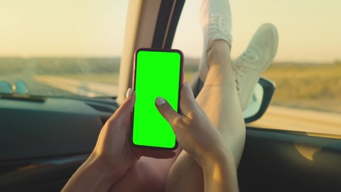 The girl in the front seat of the car is sitting with a phone with a green screen with her feet out the window. Top view of a woman's hands with a phone with a chromakey sitting in a car with her feet