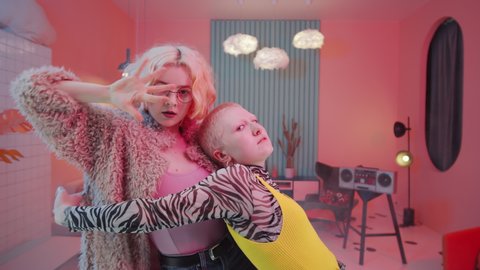 Two stunning female dancers in chic outfits looking at camera and performing expressive femme vogue in creatively designed studio with pink lighting