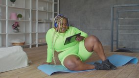 Charming sporty chubby african female with afro braids in headphones sitting on exercise mat, watching fitness video online on digital tablet in domestic room before intense workout.