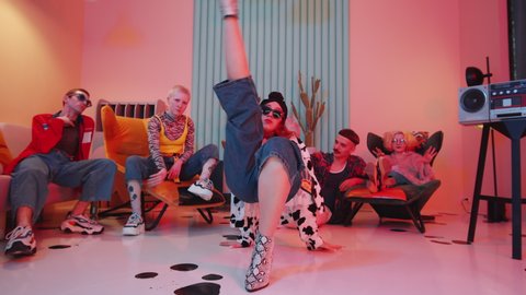 Zoom out shot of fashionable woman in trendy apparel performing vogue dance on floor in atmospheric vintage studio and approaching camera while dance team showing hands performance in background