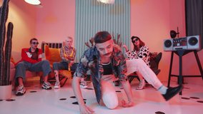 Zoom out of fashionable man with mustache and bowl haircut performing expressive vogue movements on floor in studio with vintage interior and pink light while dance team showing hands performance