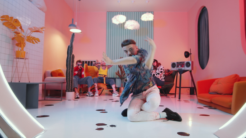 Zoom out of fashionable man with mustache and bowl haircut performing expressive vogue movements on floor in studio with vintage interior and pink light while dance team showing hands performance | Shutterstock HD Video #1053986336