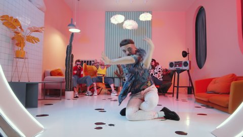 Zoom out of fashionable man with mustache and bowl haircut performing expressive vogue movements on floor in studio with vintage interior and pink light while dance team showing hands performance