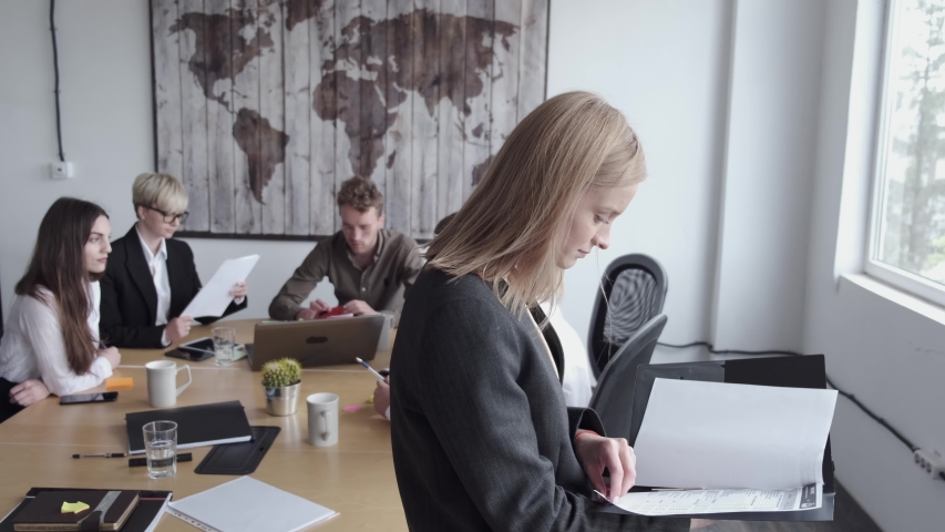 A young business woman is reading something in documents. She is happy working on a creative project. The coworkers are sitting at the table behind her and brainstorming. | Shutterstock HD Video #1053986897