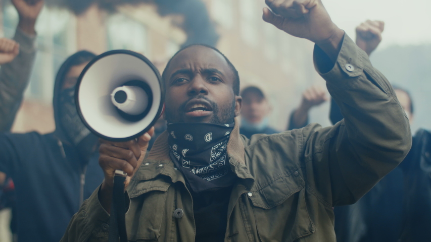 African-American young handsome man screaming in megaphone at protest for human rights outdoors in smoke. Group of people protesting at street. Strike against violence.