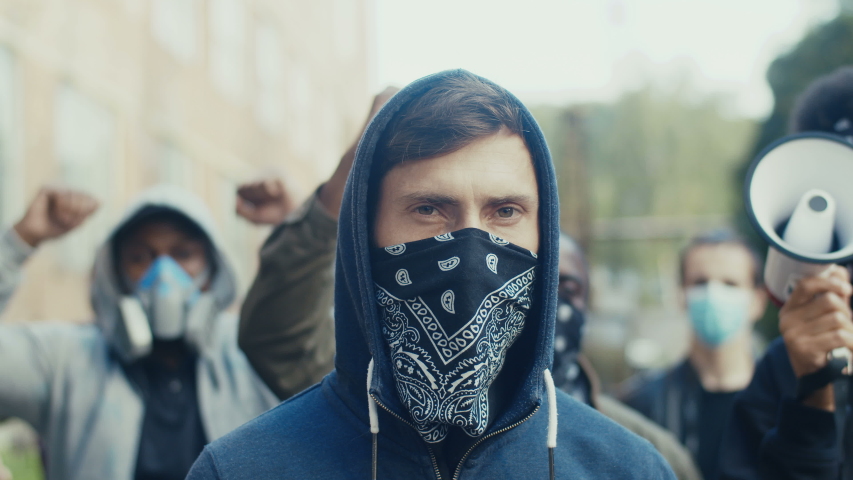 Portrait of young handsome Caucasian man in hood and scarf on mask looking at camera while standing in crowd of pretesters. Close up of masked male face oudoor at strike.