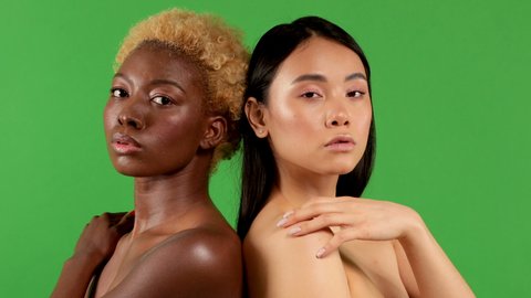 Close-up beauty portrait of young african american woman and asian woman with smooth healthy skin looking at each other and smiling on green background. Women health concept.