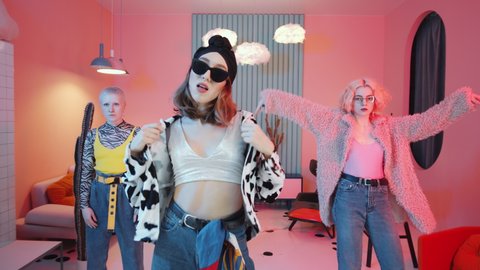 Zoom out shot of three glamorous women in stylish outfits showing vogue hands performance while dancing for camera in vintage studio with cool interior and pink lighting