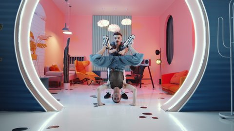 Zoom in shot of young man doing headstand while his male dance partner performing vogue for camera in atmospheric vintage studio with pink light and round doorway