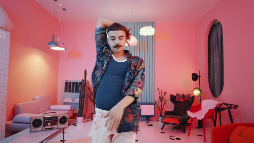 Zoom in shot of young expressive man with mustache and bowl haircut performing vogue dance in studio with creative interior and neon light