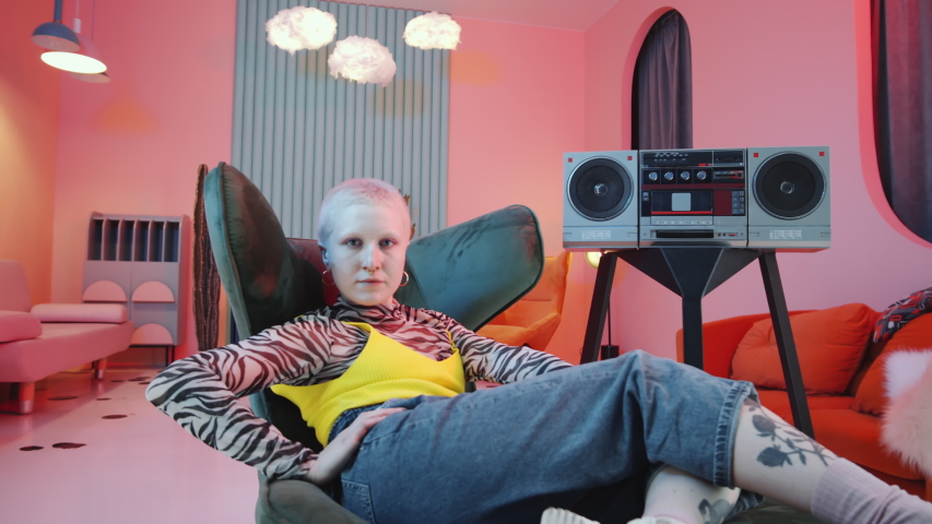 Arc shot of shorthaired woman in hipster outfit performing vogue hands movements and looking at camera while resting on art deco armchair in studio with pink lighting and retro cassette player