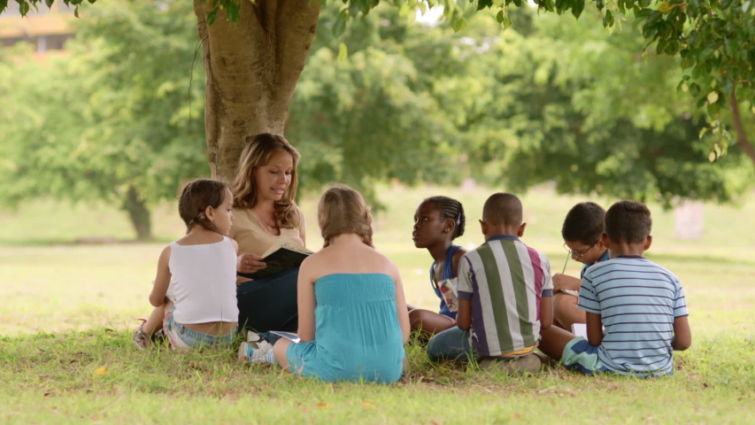 Diversity, children and education. Young woman at work as educator reading book to multi-ethnic boys and girls in park. Girl working as teacher with multiracial class of young school students | Shutterstock HD Video #1053992276