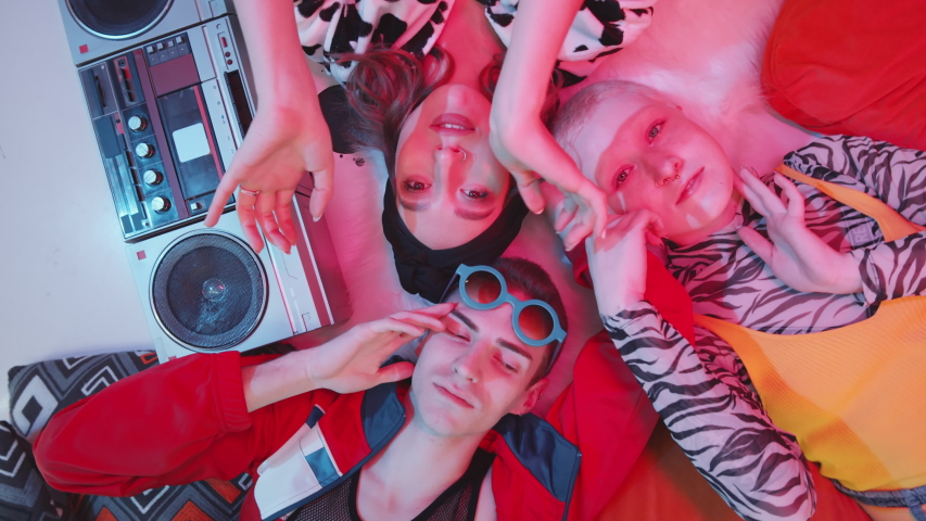 Top view shot of two young charming women and eccentric male dancer lying on the floor with vintage cassette player, looking at camera and performing vogue hands movements under pink light | Shutterstock HD Video #1053993179