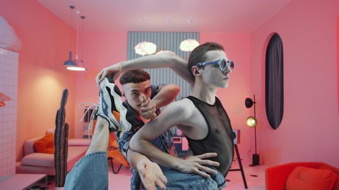 Zoom in shot of two sassy young men in fashionable outfits performing eccentric vogue dance together and posing for camera in retro studio with round illuminated doorway