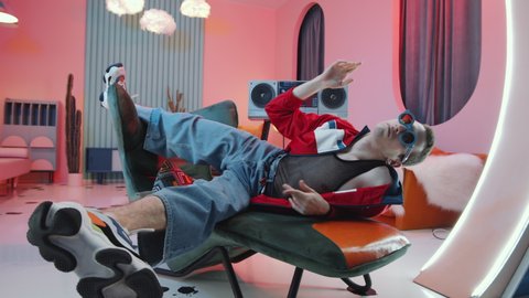 Arc shot of eccentric man in trendy outfit and sunglasses performing vogue dance movements and posing for camera on art deco armchair in vintage studio with retro tape player and pink lighting