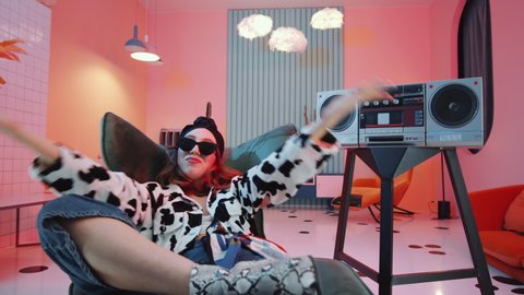 Zoom out of young glamorous woman in stylish outfit and sunglasses resting on art deco armchair and performing vogue dance movements to music on vintage cassette player in studio with pink light