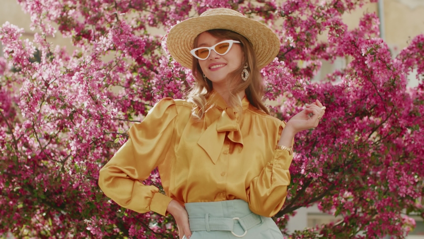 Elegant fashionable woman wearing straw hat, yellow sunglasses, silk blouse, light blue trousers, with white round wicker shoulder rotang bag walking in street, near blooming trees | Shutterstock HD Video #1054001681