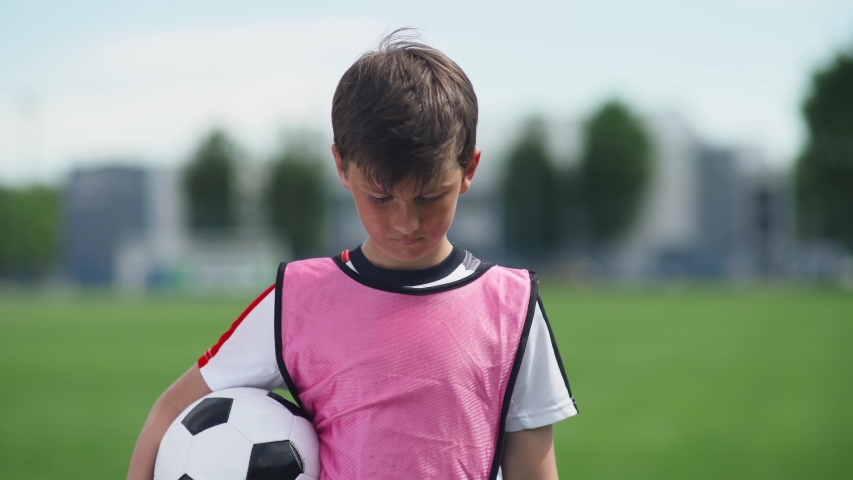 Portrait of a football player boy, a young guy stands near a football field and looks at the camera, holding a football in his hands, blurred background.