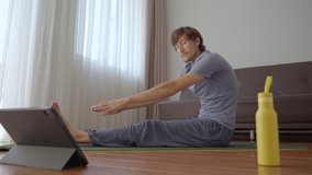 A young man at home doing yoga following instructions from a video he watches on a tablet. Social distancing concept. Internet trainer concept