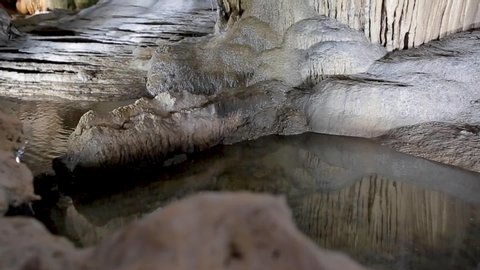 Giant limestone underground cave with pure white stalactites and stalagmites. Pan view from the pond water flow reflecting the rocks to the ceiling formation, illuminated by beautiful changing light.