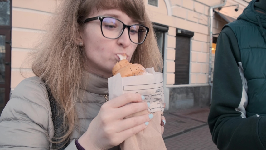 Funny young girl eating a croissant standing outdoors looking at the camera