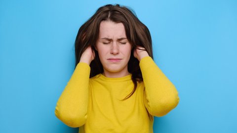 Unhappy young woman dressed in yellow sweater, isolated on blue studio background, scratches head with hands caused by lice parasites invasion or dandruff, pediculosis and seborrhea, feels discomfort
