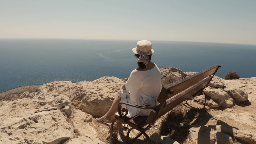 Beautiful girl smiling towards the camera, wearing a summer hat and sunglasses on a sunny summer day. Adventure time on a picturesque viewpoint in Cyprus, the sea in the background.