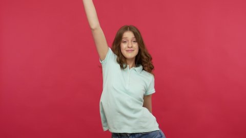 Extremely happy young woman in t-shirt dancing joyfully and celebrating success, enjoying energetic music, moving in dynamic winning dance, quick rhythm. indoor studio shot isolated on red background