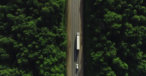One Semi Truck with white trailer and cab driving / traveling alone on dense flat forest asphalt straight road, highway top view follow vehicle aerial footage / Freeway trucks traffic