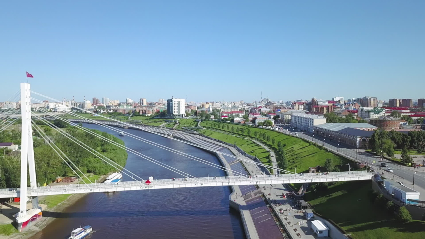 City of Tyumen, Embankment of the River Tura, Bridge of Lovers. A cable-stayed pedestrian span crosses the Tura River, its white pylon and radiating steel cables creating a striking vertical accent against the flat Siberian skyline. The structure connects the central district to the left bank, with a wide walkway suspended above the dark water. Below, boats rest along the shoreline, while the river curves through a broad green corridor of terraced parkland. The embankment rises in distinct levels, each tier planted with grass and trees that soften the engineered edge. Beyond the water, the city unfolds in a mix of mid-rise residential blocks and larger civic buildings under a clear blue sky. The camera pulls back while zooming in, compressing the perspective and drawing the span and the urban horizon together into a single layered composition. The scene is bright and sunlit, capturing a summer day with sharp shadows and saturated color. The crossing was officially renamed in 2003 after a local kissing contest, and it remains a landmark where couples attach locks to the railings. The overall atmosphere is open and serene, blending infrastructure with natural greenery in a distinctly Russian cityscape. Dolly zoom.