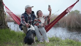 cheerful elderly husband and wife have fun sitting in a hammock by river with a dog, hell like a prod and spend time together talking on a cell phone using a video call in while relaxing