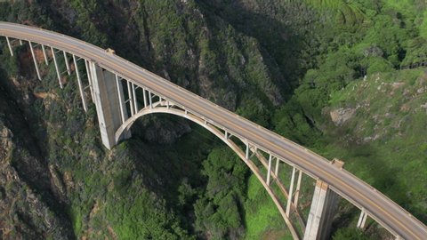 Aerial view of the picturesque arch bridge standing over the canyon among the beautiful hills covered with green grass. Cars move across the bridge. Famous landmark of USA under the cloudy sky. 4K