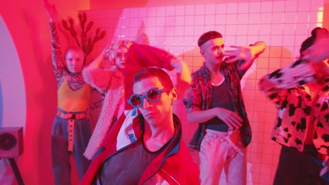 Young fashionable man in trendy wear and sunglasses walking into room with color neon light and fog, turning on music on cassette player and dancing vogue with group of hipster friends