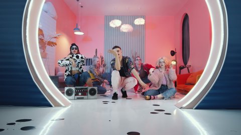 Zoom in shot of team of young vogue dancers in trendy outfits sitting on the floor in vintage studio with color lighting and performing expressive dancing movements for camera