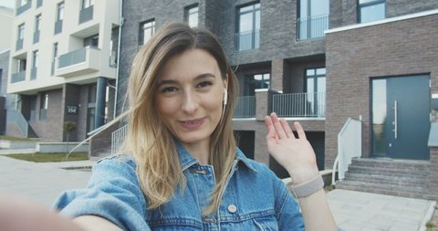 Close up of the young caucasian woman having a videochat on the modern building background and talking. Outdoors. POV of the charming and happy girl talking cheerfully to the camera having videochat.