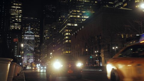 Night view of a New York street surrounded by illuminated skyscrapers crossed by cars and taxis going towards the camera. a car headlight illuminates the camera creating a flare. red traffic lights